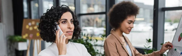 Femme d'affaires musulmane parlant sur un téléphone portable près d'un collègue afro-américain et tableau à feuilles mobiles dans le bureau, bannière — Photo de stock