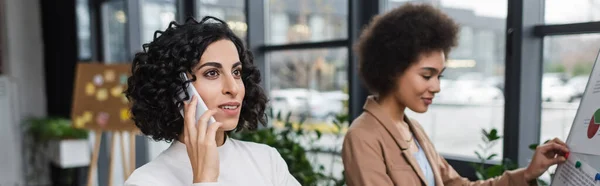 Donna d'affari musulmana che parla al cellulare vicino al collega afro-americano e lavagna a fogli mobili in ufficio, banner — Foto stock