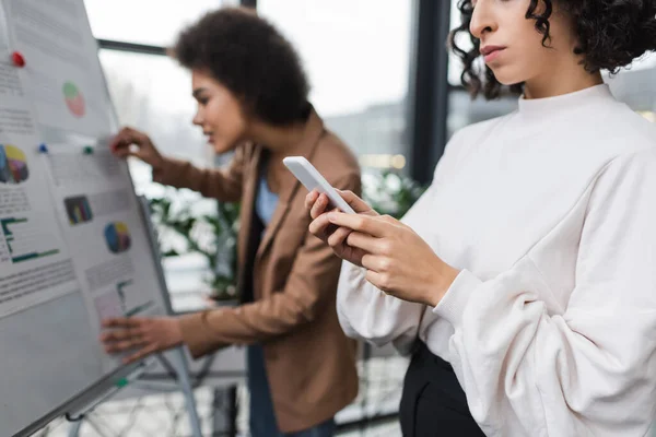 Empresaria musulmana usando teléfono celular cerca borrosa colega afroamericano y rotafolio - foto de stock