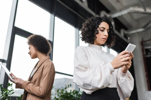 Vista de ángulo bajo de la mujer de negocios árabe utilizando el teléfono móvil cerca de un colega afroamericano con tableta digital en la oficina - foto de stock