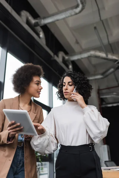 Empresaria musulmana hablando en smartphone y usando tableta digital cerca de colega afroamericano en la oficina - foto de stock