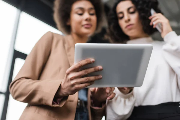 Low angle view of digital tablet in hand of blurred african american businesswoman near arabian colleague talking on cellphone — Fotografia de Stock
