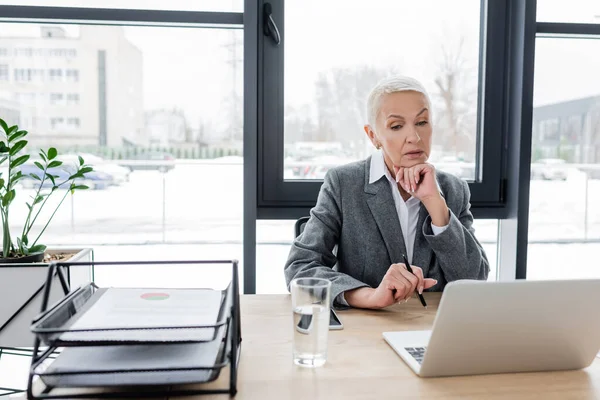 Thoughtful businesswoman looking at laptop near glass of water on desk — Stockfoto