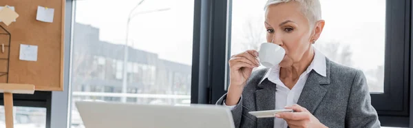 Economista senior beber café cerca de ordenador portátil borrosa en la oficina, pancarta - foto de stock