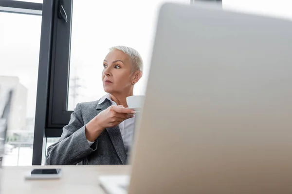 Thoughtful businesswoman with coffee cup looking away near blurred laptop — Photo de stock
