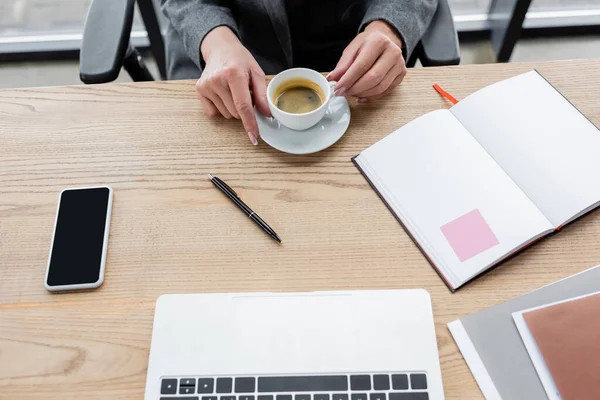 Vista parcial del financiero cerca de la taza de café, portátil vacío, computadora portátil y teléfono inteligente con pantalla en blanco - foto de stock