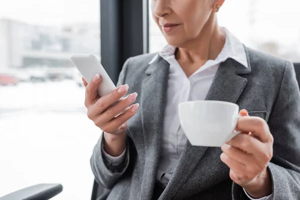 Vista parziale del banchiere con tazza di caffè utilizzando il telefono cellulare in ufficio — Foto stock