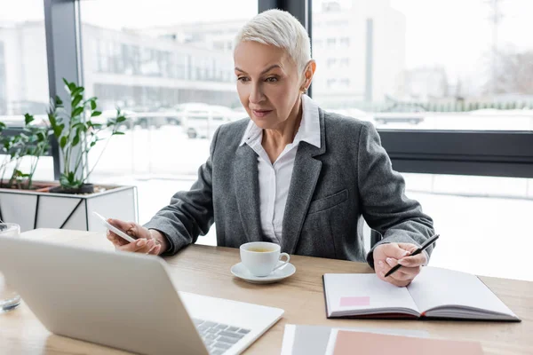 Surprised banker with smartphone and pen looking at laptop during video call — Stock Photo