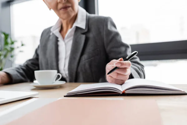 Cropped view of blurred economist with pen sitting near notebook and coffee cup - foto de stock