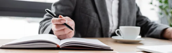 Cropped view of banker holding pen near notebook while sitting near coffee cup on blurred background, banner — Stock Photo