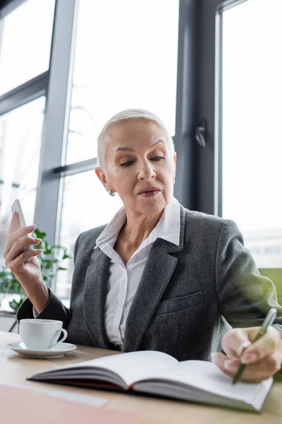 Banquier senior tenant smartphone près d'une tasse de café et écrivant dans un carnet flou — Photo de stock