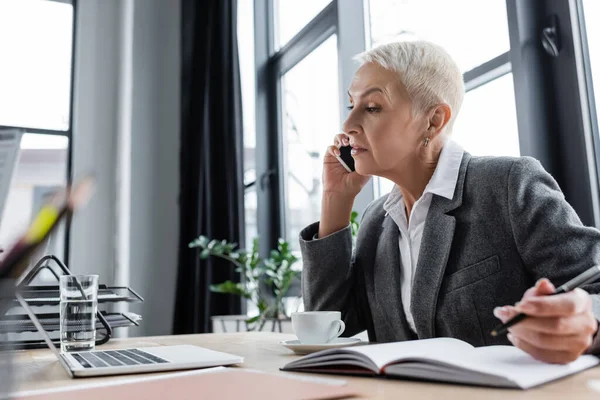 Businesswoman talking on smartphone while looking at laptop near notebook — стоковое фото