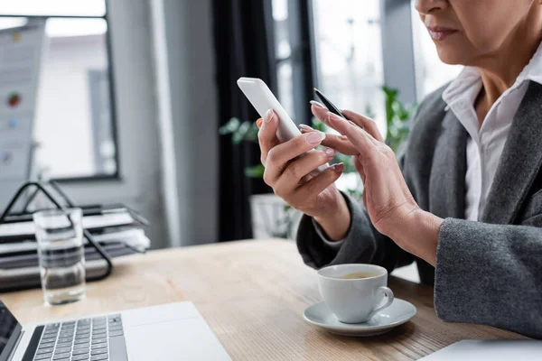 Vista ritagliata del banchiere utilizzando smartphone vicino laptop e tazza di caffè sulla scrivania — Foto stock