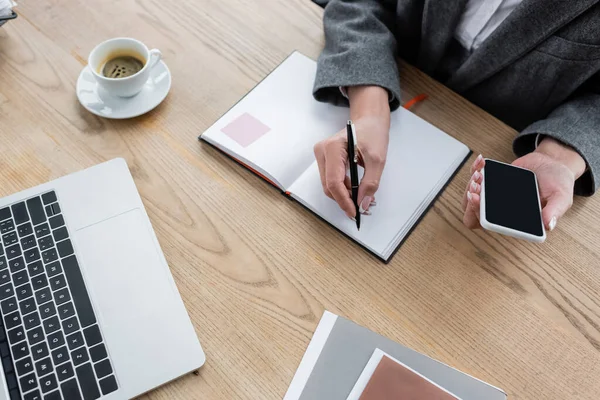 Vue recadrée du banquier tenant smartphone avec écran vierge et écriture dans un ordinateur portable — Photo de stock