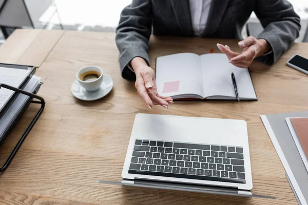 Teilbild eines Bankers, der während einer Videokonferenz auf Laptop neben leerem Notizbuch und Kaffeetasse gestikuliert — Stockfoto