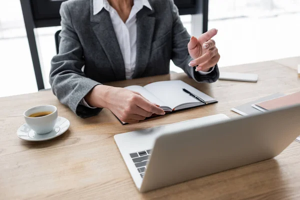 Ausgeschnittene Ansicht des Bankers, der während eines Videogesprächs mit dem Finger auf Laptop in der Nähe von Notebook und Kaffeetasse zeigt — Stockfoto