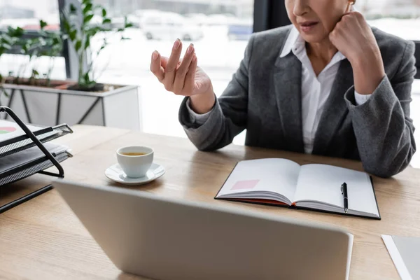 Vue recadrée de geste de l'économiste lors de la conférence en ligne sur ordinateur portable près ordinateur portable vierge et tasse de café — Photo de stock