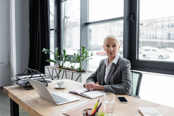 Stilvoller Banker sitzt am Arbeitsplatz in der Nähe von Geräten und Notebooks und blickt in die Kamera — Stockfoto