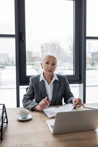 Thoughtful economist holding pen while sitting near notebook and laptop — Stock Photo