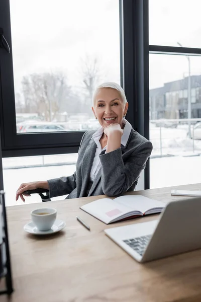 Successful banker smiling at camera near blank notebook, coffee cup and blurred laptop — Stock Photo