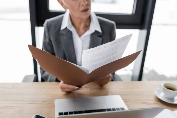 Partial view of blurred banker holding folder with documents near laptop and coffee cup — Foto stock