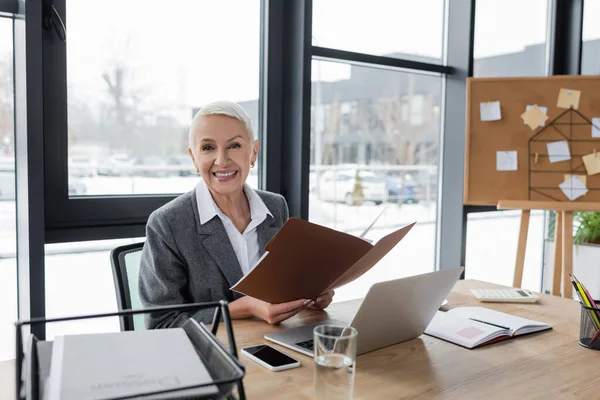 Happy banker with folder smiling at camera near laptop and cellphone with blank screen — Stock Photo