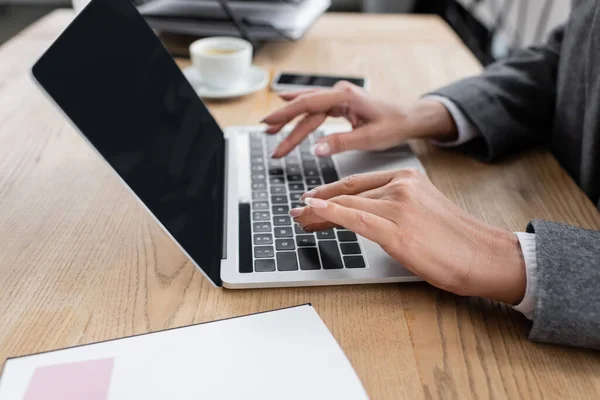 Vista ritagliata della donna d'affari che digita sul computer portatile con schermo vuoto vicino alla tazza di caffè offuscata — Foto stock