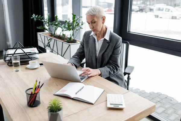 Senior Buchhalter tippt auf Laptop neben leerem Notizbuch und Taschenrechner — Stockfoto