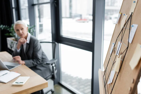 Blurred banker sitting at workplace and looking at corkboard with paper notes - foto de stock