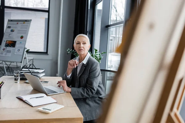 Senior-Geschäftsfrau schaut auf verschwommenes Flipchart, während sie am Arbeitsplatz neben Laptop sitzt — Stockfoto