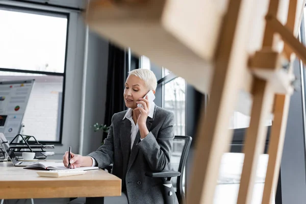 Banker talking on cellphone and writing in notebook on blurred foreground - foto de stock