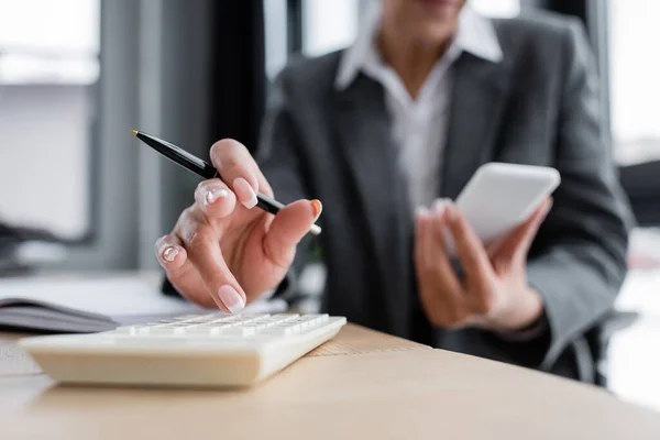 Partial view of accountant working on calculator and holding smartphone on blurred background - foto de stock