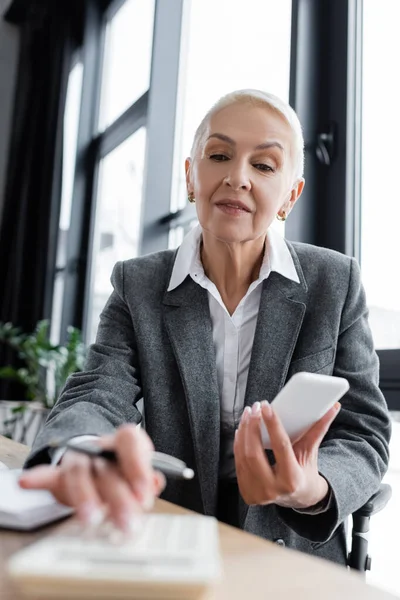 Accountant holding mobile phone while using blurred calculator in office - foto de stock