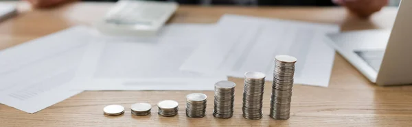 Piles de pièces d'argent près d'un ordinateur portable flou et des documents sur le bureau, bannière — Photo de stock