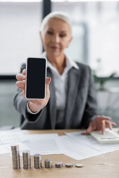 Banker showing smartphone with blank screen near silver coins and documents on blurred background - foto de stock