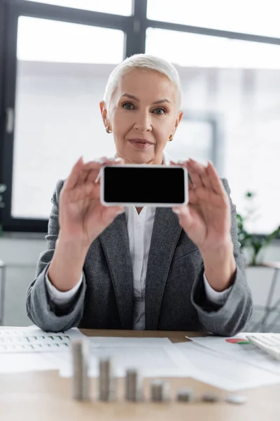 Positive banker holding mobile phone with blank screen while looking at camera near blurred documents - foto de stock