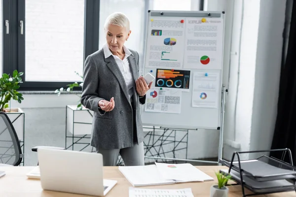 Banker with cellphone pointing at laptop during video call near graphs on flip chart — Foto stock