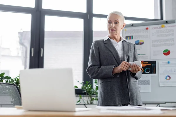 Senior Banker mit Smartphone schaut während Online-Konferenz auf Laptop und fast verschwommenes Flipchart — Stockfoto