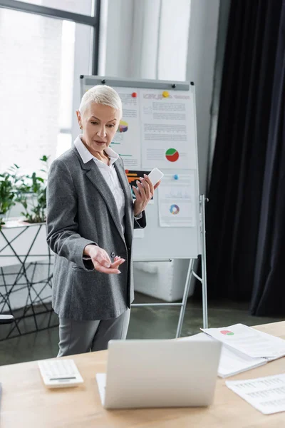 Leitender Banker mit Handy, das während Videokonferenz auf Laptop zeigt, in der Nähe von verschwommenem Flipchart — Stockfoto