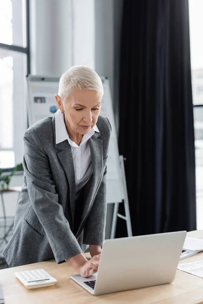 Senior banker in formal wear standing at workplace and using laptop — Stock Photo