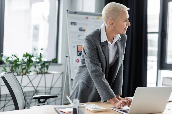 Stylish economist standing at workplace, using laptop and looking away — Photo de stock