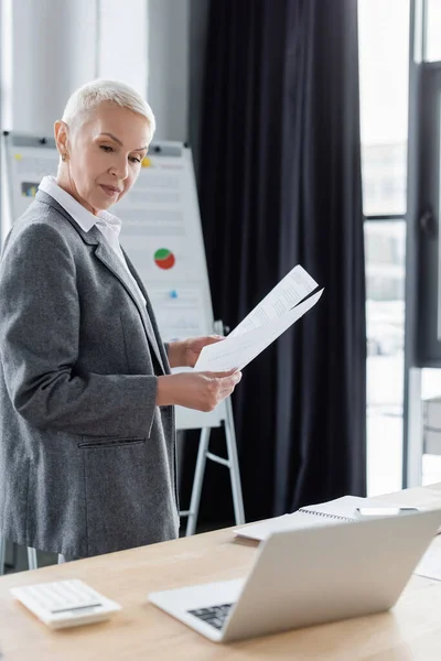 Стильна бізнес-леді стоїть з документами на робочому місці під час онлайн-конференції на ноутбуці — стокове фото