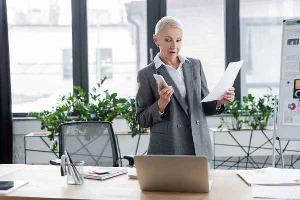 Banker with smartphone and document looking at laptop during video conference in office — Stock Photo