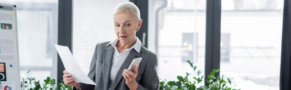 Surprised banker holding paper and looking at mobile phone in office, banner — Stock Photo