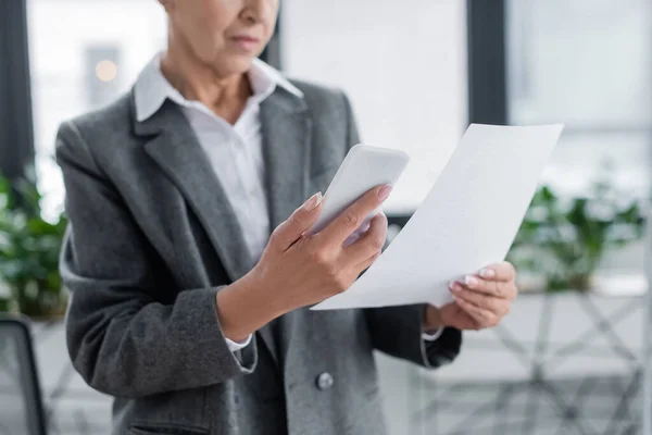 Partial view of businesswoman holding mobile phone and paper on blurred background — Stock Photo