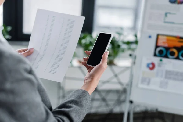 Partial view of banker holding smartphone and document with analytics near blurred flip chart — Stock Photo