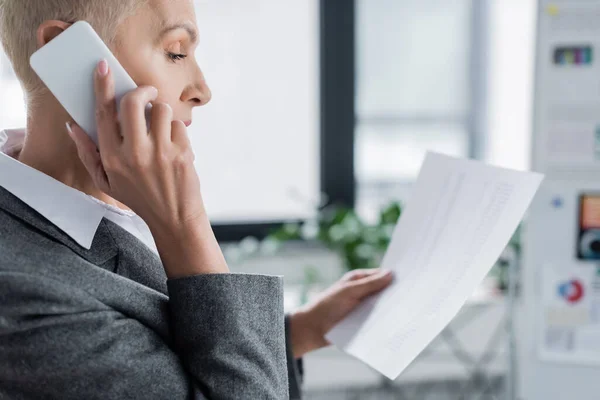 Side view of senior economist holding document and talking on cellphone in office — Stock Photo