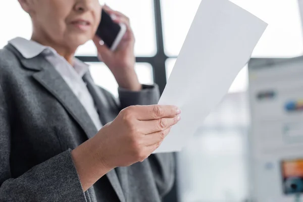 Selective focus of white paper in hand of cropped banker talking on smartphone on blurred background — Stock Photo