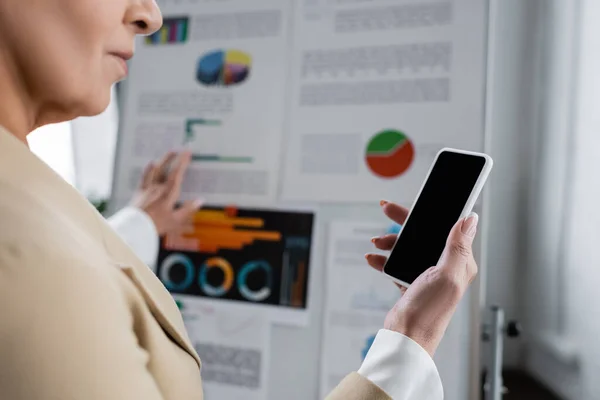 Ausgeschnittene Ansicht eines Bankers, der sein Smartphone mit leerem Bildschirm in der Nähe eines unscharfen Flipcharts mit Grafiken hält — Stockfoto