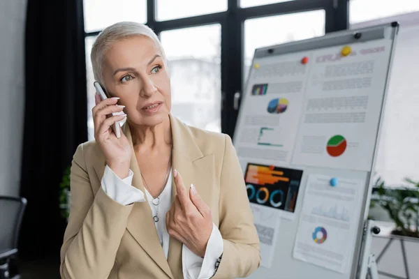 Stylish businesswoman talking on mobile phone near flip chart on blurred background — Stock Photo